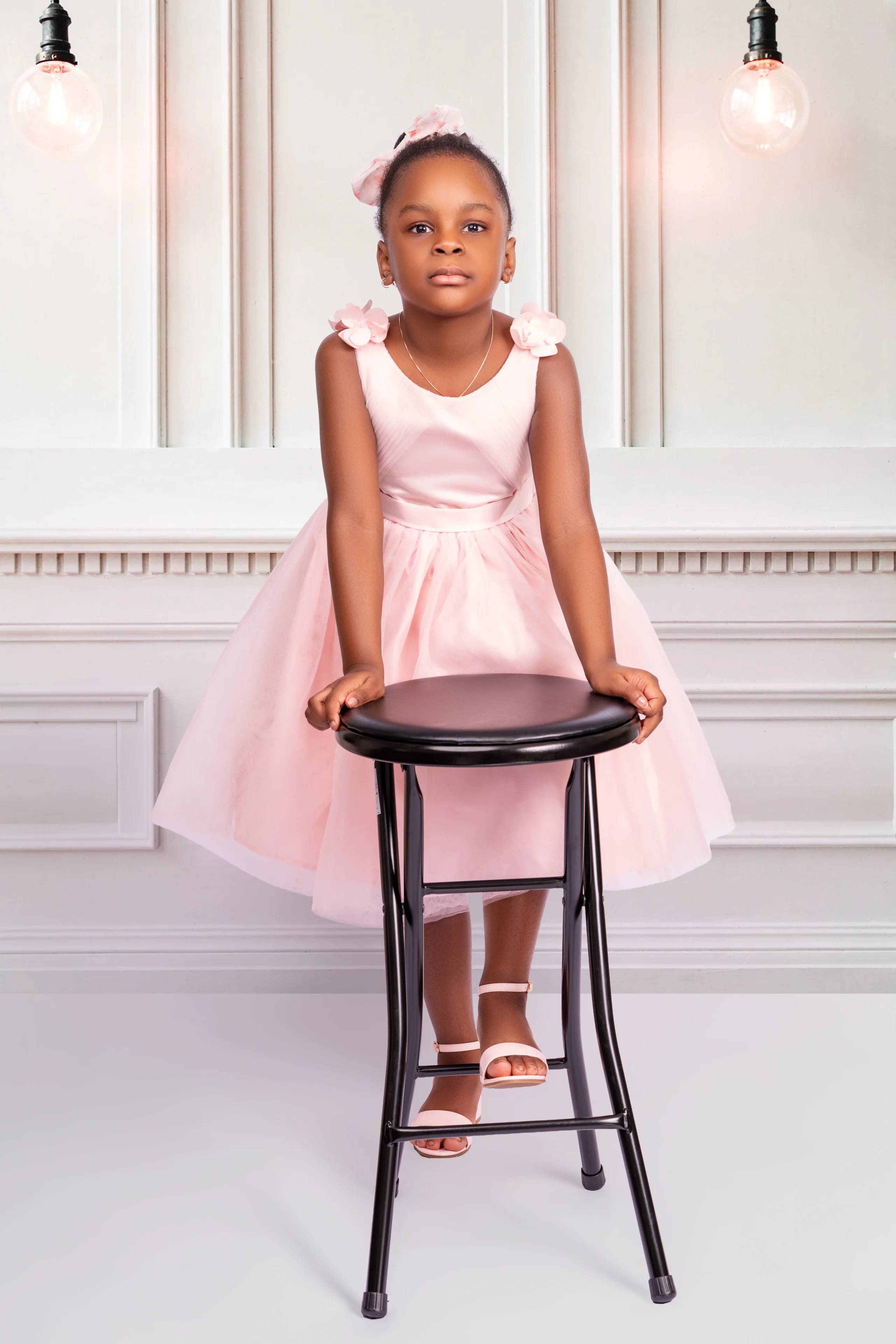 Girl in a pink dress behind a stool under hanging filament bulbs