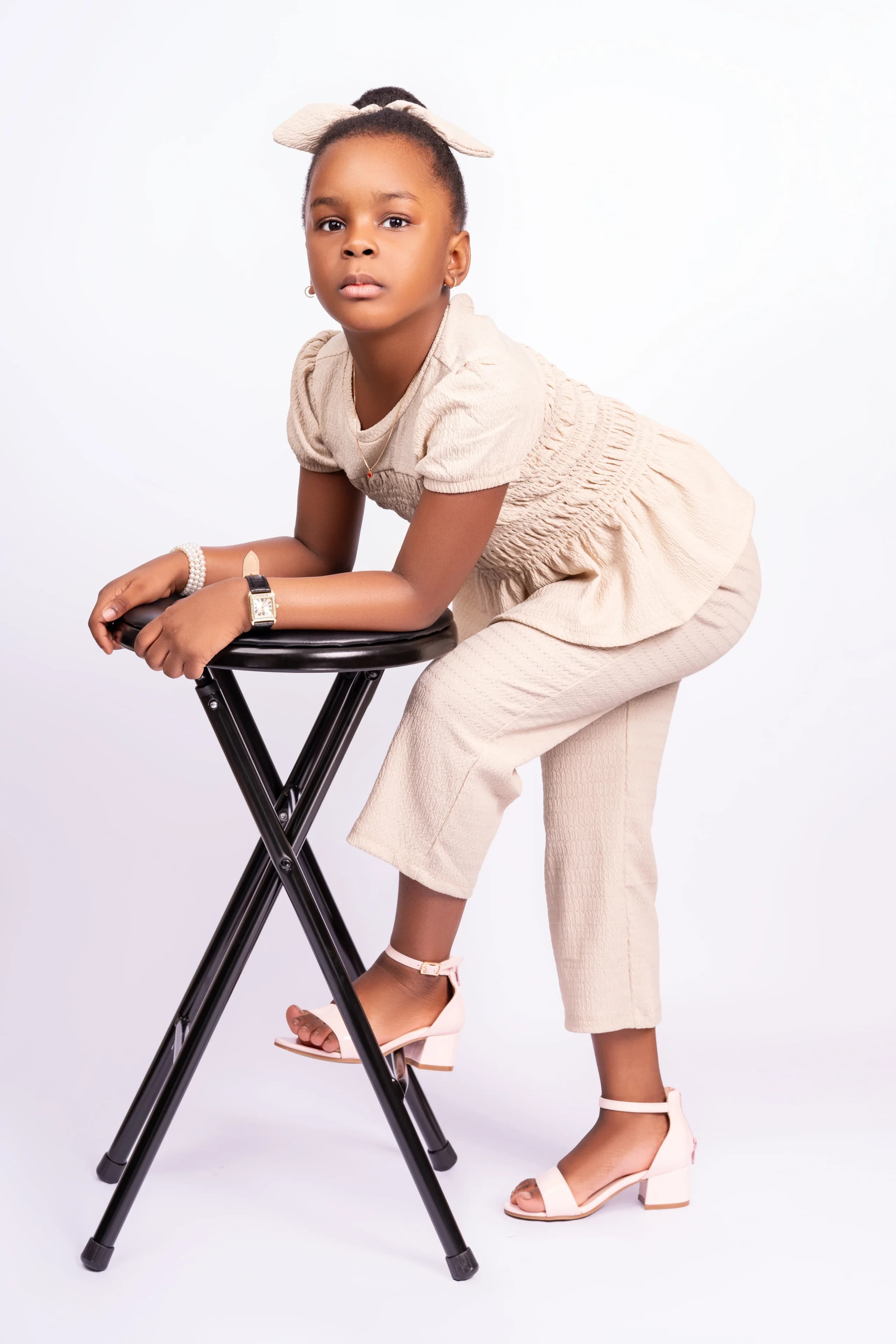 Girl in a beige outfit leaning on a stool in a white studio